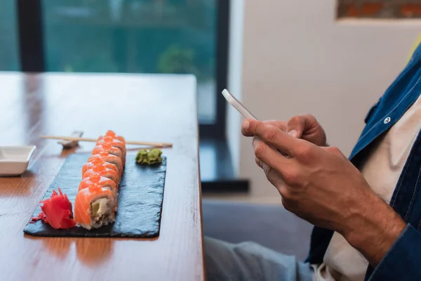 Vista recortada del hombre charlando en el teléfono móvil cerca de rollos de sushi en la mesa del restaurante - foto de stock