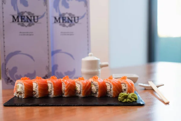 Tasty sushi rolls with red caviar and wasabi near blurred menu brochures on wooden table — Fotografia de Stock