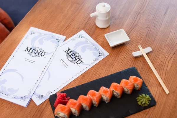 Brochures de menu à proximité de délicieux rouleaux de sushi, baguettes, casserole de soja et bol sur table en bois — Photo de stock