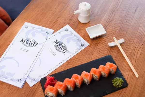 Folletos de menú cerca de deliciosos rollos de sushi, palillos, olla de salsa de soja y tazón en mesa de madera - foto de stock