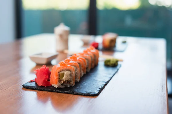 Deliciosos rollos de sushi cerca de jengibre en plato negro y mesa de madera, fondo borroso - foto de stock