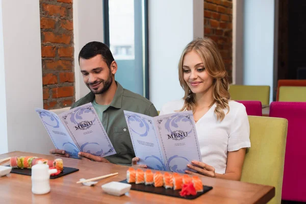 Happy couple choosing meal from menu near blurred sushi rolls in restaurant — Fotografia de Stock