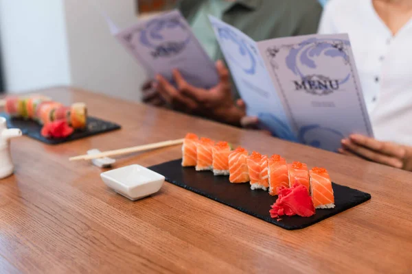 Vue recadrée du couple flou avec des brochures de menu près de savoureux rouleaux de sushi — Photo de stock