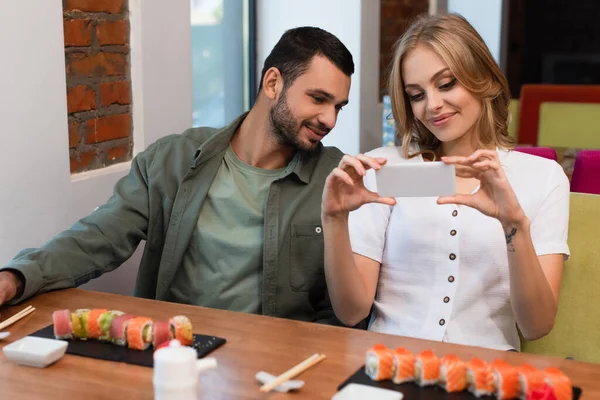 Mulher sorrindo tirar fotos de rolos de sushi durante o jantar com o namorado — Fotografia de Stock
