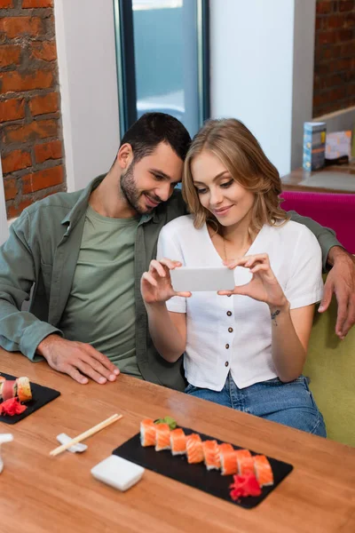 Woman taking photo of sushi rolls on smartphone near smiling boyfriend — Foto stock