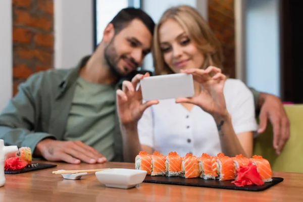 Donna sfocata scattare foto di rotoli di sushi vicino fidanzato felice in sushi bar — Foto stock