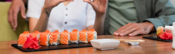 Partial view of blurred couple near set of tasty sushi rolls and sauce bowl, banner — Stock Photo