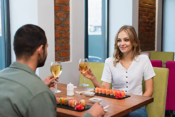 Jovem casal segurando copos de vinho branco perto de deliciosos rolos de sushi no restaurante — Fotografia de Stock