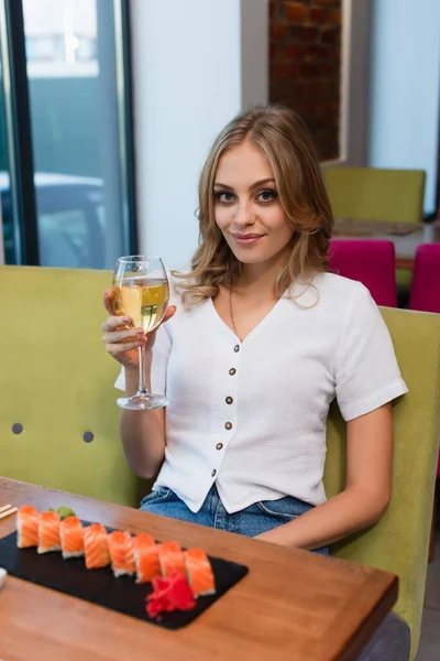 Femme heureuse avec un verre de vin blanc regardant la caméra près des rouleaux de sushi flous — Photo de stock