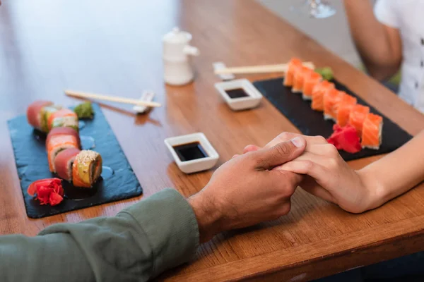 Vista cortada do casal de mãos dadas perto de rolos de sushi e tigelas com molho de soja na mesa — Fotografia de Stock