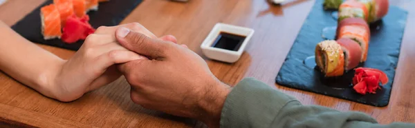 Partial view of couple holding hands near sets of sushi rolls and bowl with soy sauce, banner — Fotografia de Stock