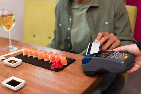 Vista cortada de homem desfocado pagando através de leitor de cartão de crédito perto de rolos de sushi e copo de vinho — Fotografia de Stock