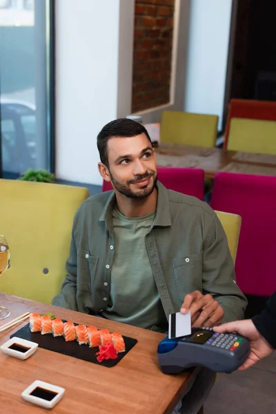 Homme souriant payer par carte de crédit via terminal de paiement près de savoureux sushi — Photo de stock