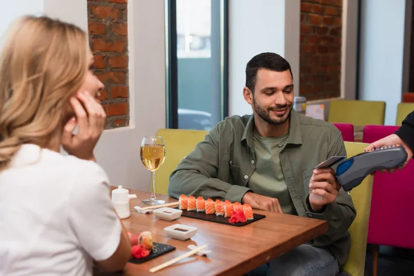 Blurred woman near boyfriend paying for dinner in sushi bar through credit card reader — Stock Photo