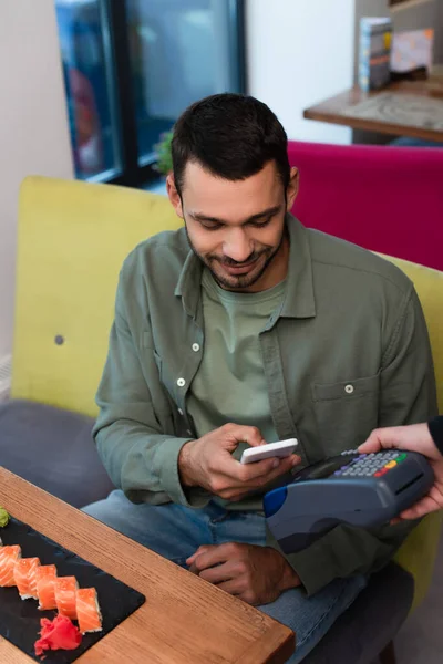 Lächelnder Mann hält Smartphone in der Nähe von Zahlungsterminal und Teller mit Sushi-Rollen — Stockfoto