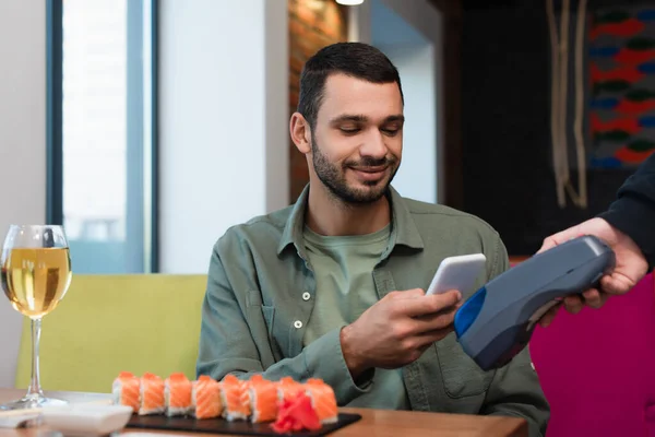 Young man paying with smartphone through payment terminal for tasty sushi and glass of white wine — Stock Photo