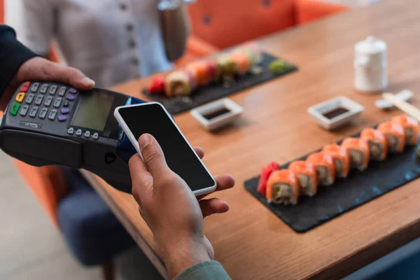 Ausgeschnittene Ansicht eines Mannes mit Smartphone mit leerem Bildschirm in der Nähe von Zahlungsterminal und Tisch mit Sushi-Brötchen — Stockfoto