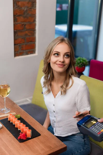 Mulher sorridente com cartão de crédito pagando através do terminal para rolos de sushi e copo de vinho — Fotografia de Stock
