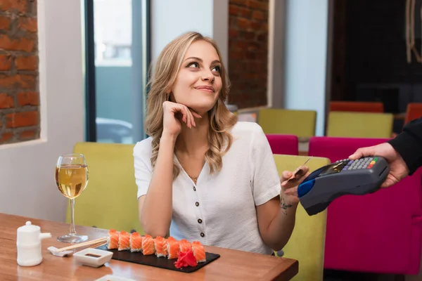 Waiter holding payment terminal near woman with credit card, sushi rolls and glass of white wine — Stock Photo