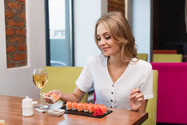 Mujer alegre con palillos sosteniendo rollo de sushi cerca de salsa de soja y copa de vino blanco - foto de stock