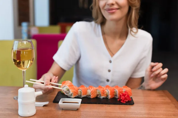 Vista recortada de la mujer borrosa sonriendo mientras sostiene sabroso rollo de sushi cerca de un vaso de vino blanco - foto de stock