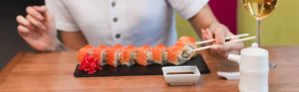 Vue partielle de la femme avec baguettes près des rouleaux de sushi, sauce soja et verre de vin blanc, bannière — Photo de stock