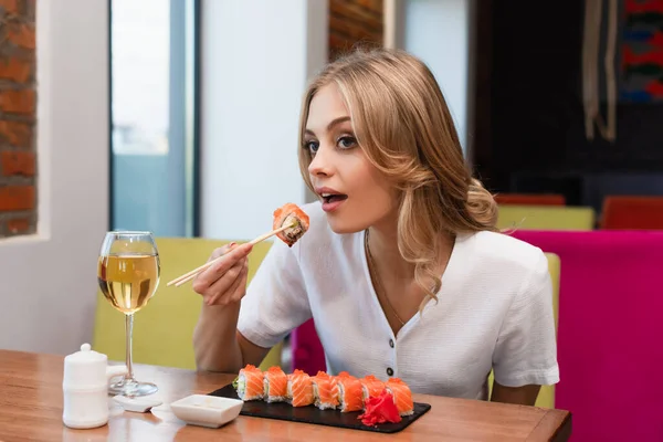 Mulher loira comendo sushi saboroso perto de vidro ou vinho branco — Fotografia de Stock