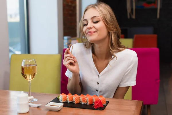 Joyful woman with chopsticks near plate with sushi, glass of white wine and soy sauce — Foto stock