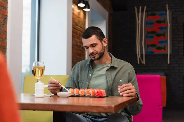 Young man holding chopsticks near sushi and glass of red wine, blurred foreground — Foto stock