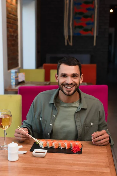 Hombre feliz con palillos mirando a la cámara cerca de sushi sabroso - foto de stock