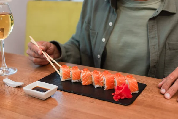 Vista cortada do homem com pauzinhos perto de delicioso sushi, tigela com molho de soja e copo de vinho tinto — Fotografia de Stock