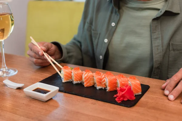 Cropped view of man with chopsticks near delicious sushi, bowl with soy sauce and glass of red wine — Fotografia de Stock