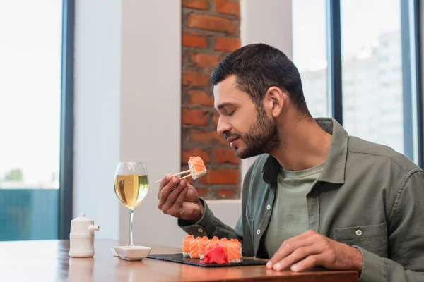 Young man with closed eyes holding delicious sushi with chopsticks near glass or white wine — Foto stock