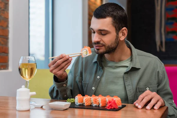 Young man holding tasty sushi with chopsticks near glass of wine on table — Stock Photo