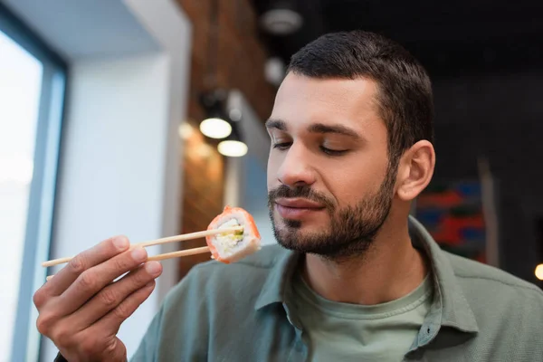 Jovem barbudo segurando sushi rolo com pauzinhos no restaurante — Fotografia de Stock