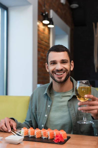 Hombre feliz con copa de vino blanco mirando a la cámara cerca del plato con sushi delicioso - foto de stock