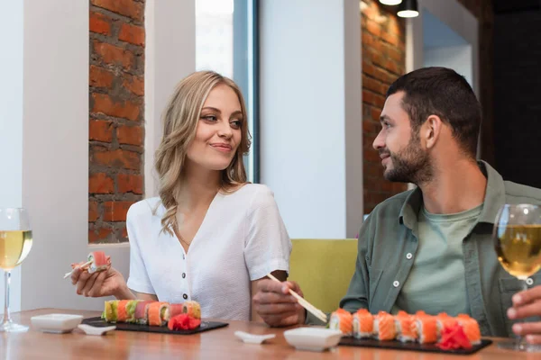 Casal sorrindo olhando um para o outro perto de deliciosos rolos de sushi e copos de vinho branco — Fotografia de Stock