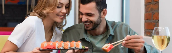 Happy woman showing set of delicious sushi to smiling man in restaurant, banner — Stock Photo