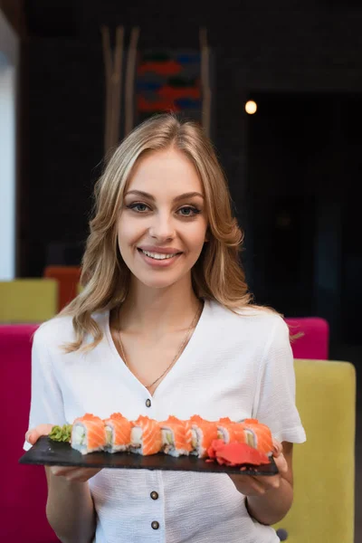 Mujer rubia feliz mirando a la cámara mientras sostiene sabrosos rollos de sushi - foto de stock