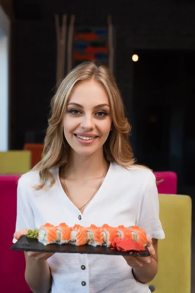 Счастливая блондинка смотрит в камеру, держа в руках вкусные суши-роллы — стоковое фото