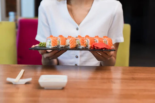 Vue partielle de la femme tenant la plaque de rouleaux de sushi près de baguettes floues et bol à sauce — Photo de stock