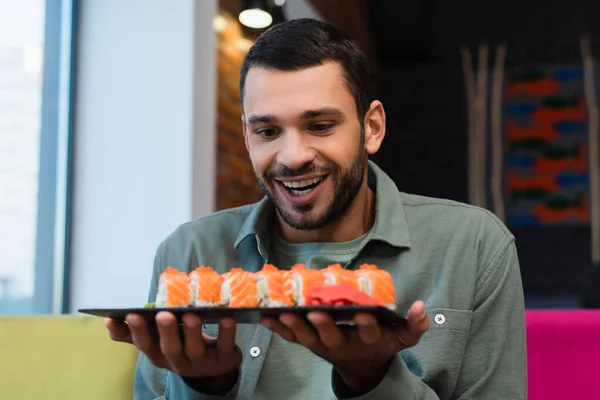 Homem espantado segurando prato com conjunto de saborosos rolos de sushi no restaurante — Fotografia de Stock