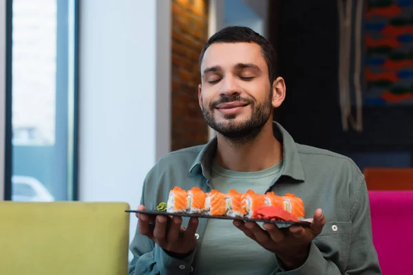 Homem satisfeito com olhos fechados segurando prato com saborosos rolos de sushi no restaurante — Fotografia de Stock