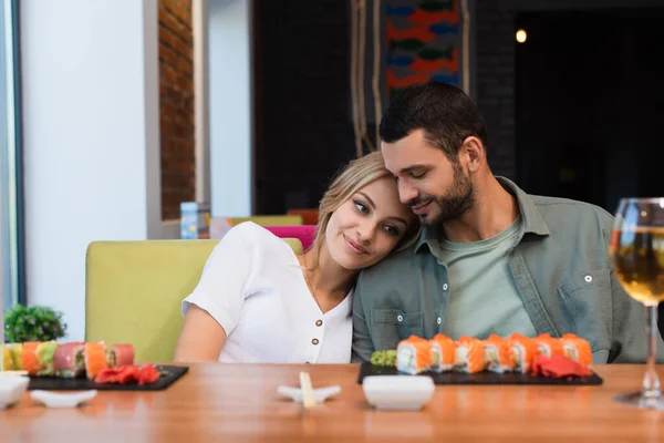 Joven y feliz pareja sentado cerca borrosa sushi rollos y copa de vino blanco en el restaurante - foto de stock