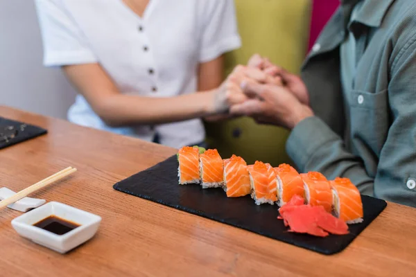 Vista cortada de casal desfocado de mãos dadas perto da placa com deliciosos rolos de sushi no restaurante — Fotografia de Stock