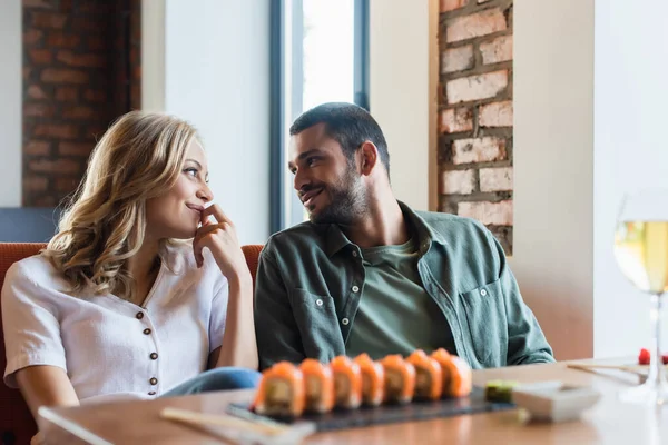 Coppia gioiosa sorridente a vicenda vicino involtini di sushi sfocati e bicchiere di vino bianco — Foto stock