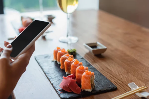 Visão parcial da mulher segurando telefone celular com tela em branco perto de rolos de sushi e vidro borrado de vinho tinto — Fotografia de Stock