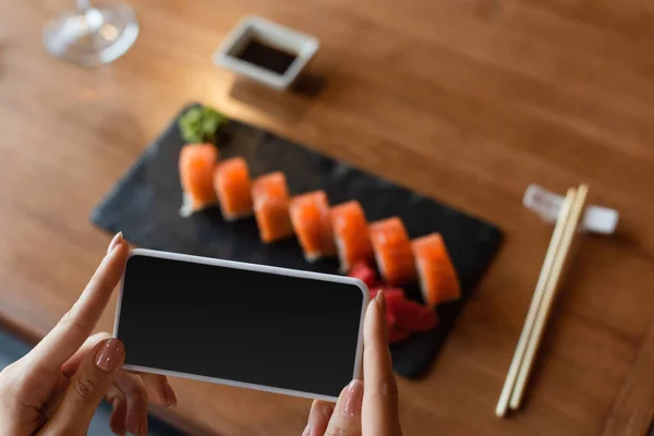 Partial view of woman taking photo of delicious sushi on mobile phone, blurred background — Fotografia de Stock