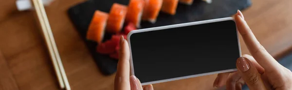 Vista cortada de mulher segurando smartphone com tela em branco perto de rolos de sushi borrados, banner — Fotografia de Stock
