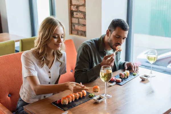 Mujer feliz comiendo sabrosos rollos de sushi cerca de novio y vasos de vino blanco - foto de stock